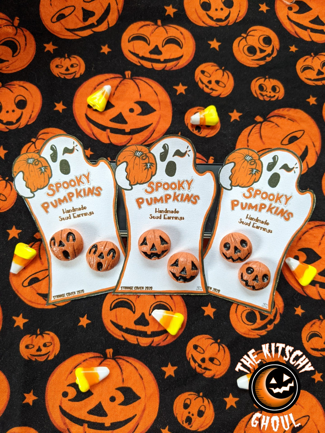 3 pairs of Hand sculpted Spooky Pumpkin Earrings on their ghost shaped cards, among candy corn on a vintage pumpkin print fabric background